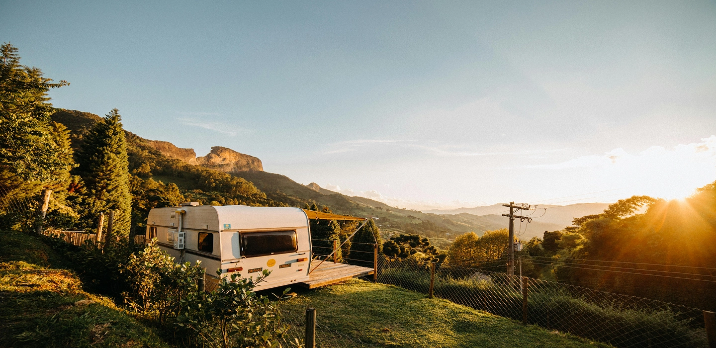 A white camper van parked on a grassy hillside overlooks mountains and a valley at sunset, with trees and a wire fence nearby.