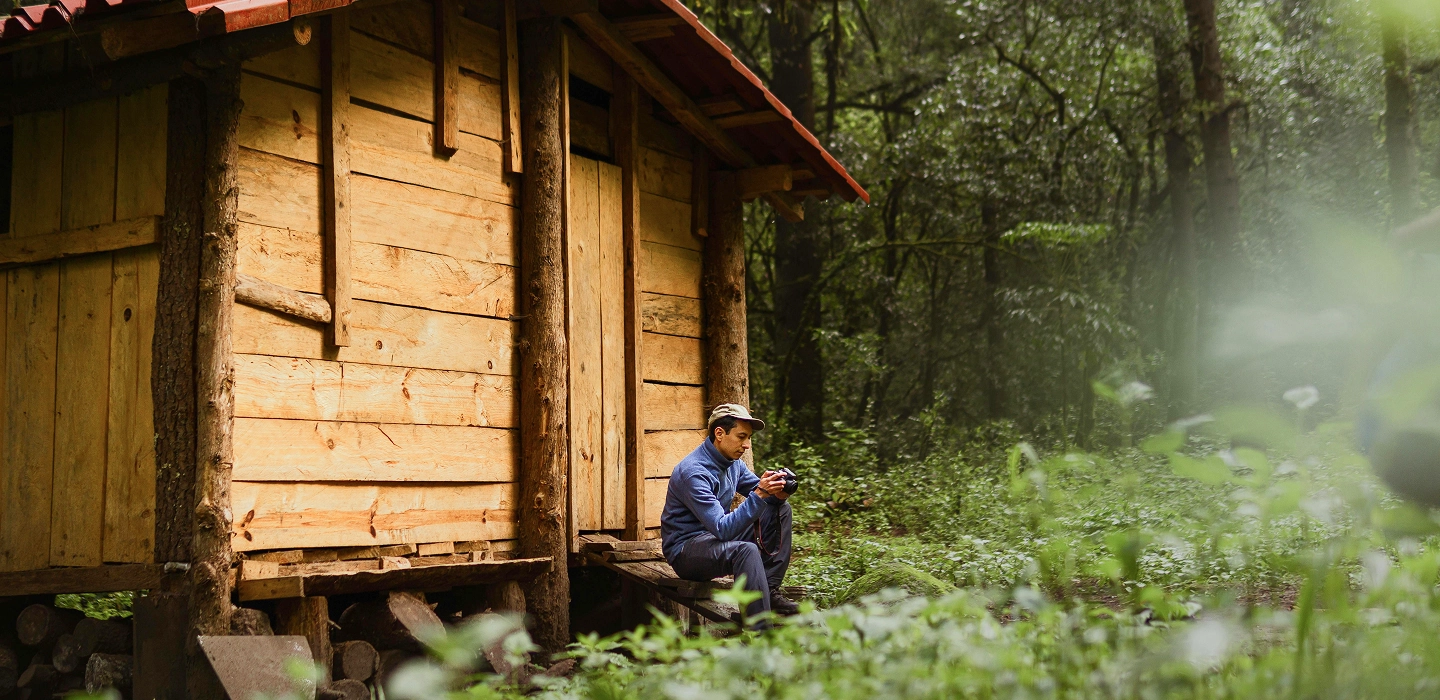 A person sits on the steps of a wooden cabin surrounded by dense green forest, holding a mug and looking down.