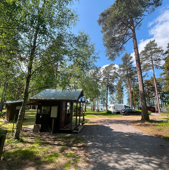 A small wooden cabin stands among tall trees near a gravel path, with parked vehicles and a white caravan visible in the background under a partly cloudy sky.