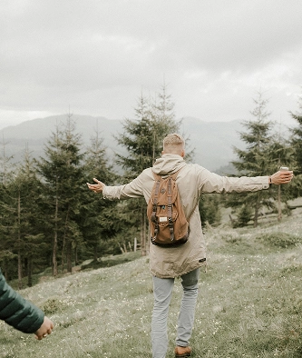 A person with a rucksack walks on a grassy hillside with arms outstretched, surrounded by pine trees and mountains in the background.