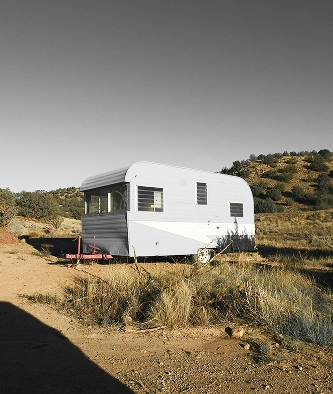 A white vintage caravan is parked on a patch of dry grass and dirt, with hills and sparse vegetation in the background under a clear sky.