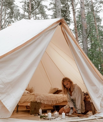 A woman sits on the floor at the entrance of a canvas tent in a forest, with a bed and various items visible inside the tent.