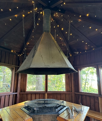 A large metal fireplace hood hangs above a grill in a wooden gazebo with fairy lights on the ceiling and windows showing trees outside.