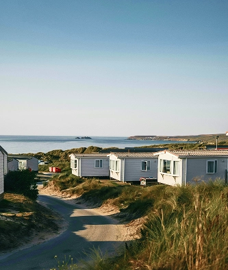 Row of caravans along a sandy path near the coast, with grassy dunes and calm sea visible under a clear blue sky.