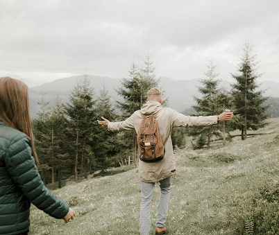 Two people walk outdoors on a grassy hill surrounded by pine trees, with one person ahead wearing a rucksack and stretching out their arms.