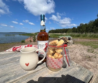 A ceramic mug, a jar of biscuits, and a bottle of Oban whisky sit on a wooden table outdoors by a lake, with trees and blue sky in the background.