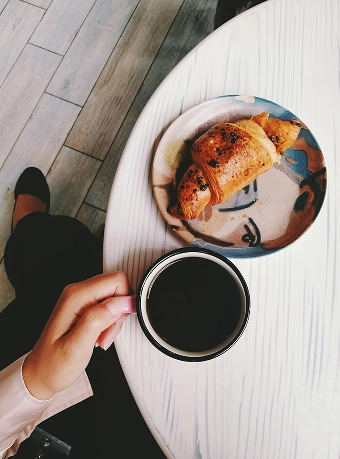 A person holds a cup of black coffee next to a plate with a croissant on a white table.