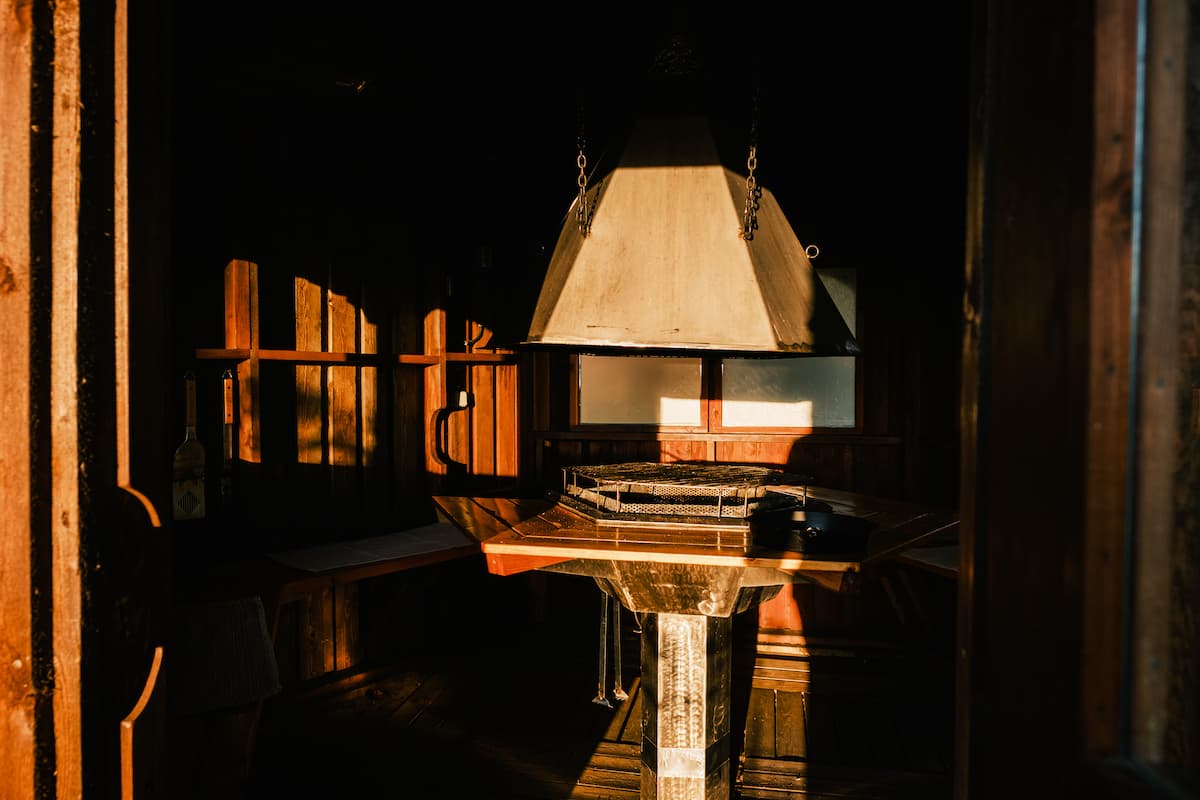 Sunlight shines on a central grille inside a wooden room, highlighting a metal hood above and creating warm shadows on the walls.