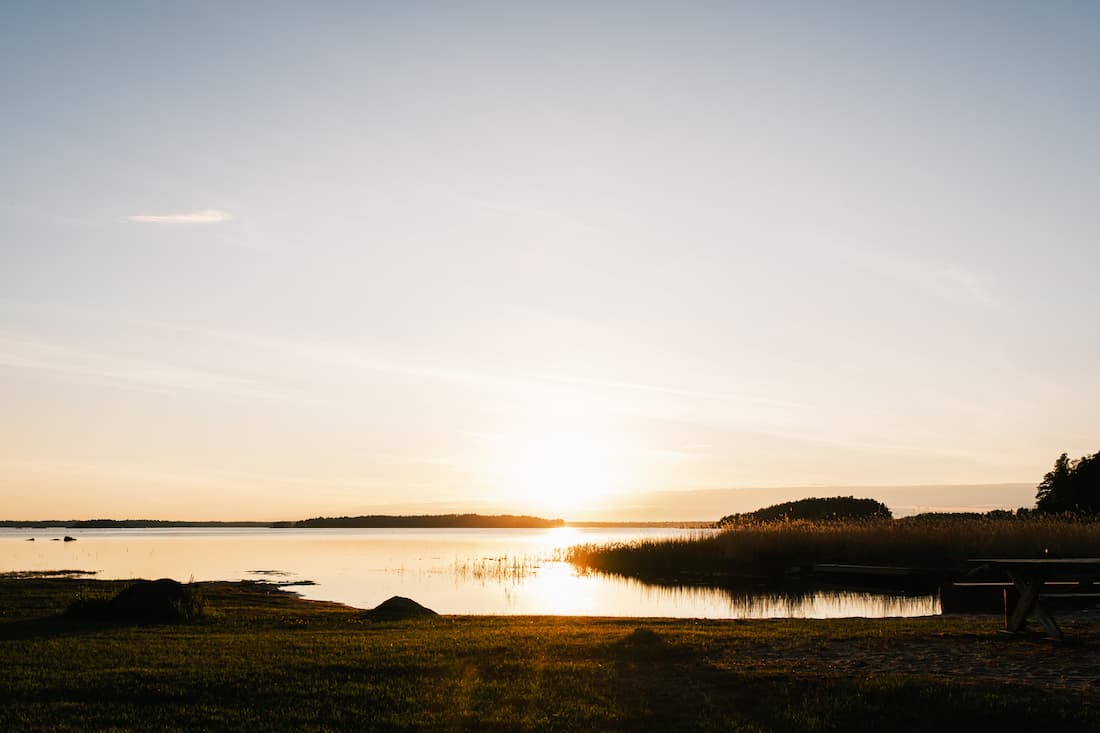 Sunset over a calm lake with grassy banks, distant trees, and a clear sky. A wooden bench is visible on the right side of the image.