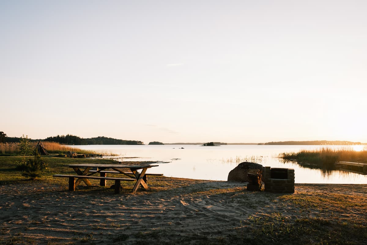 A wooden picnic table and fire pit sit on sandy ground by a calm lakeside at sunset, with reeds and trees in the background.