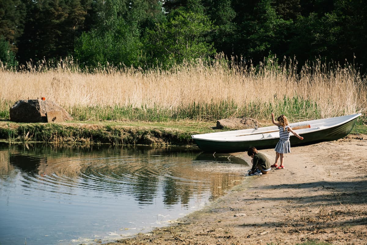 Two children play by the water's edge near a rowing boat on a sandy shore, with tall grass and trees in the background.