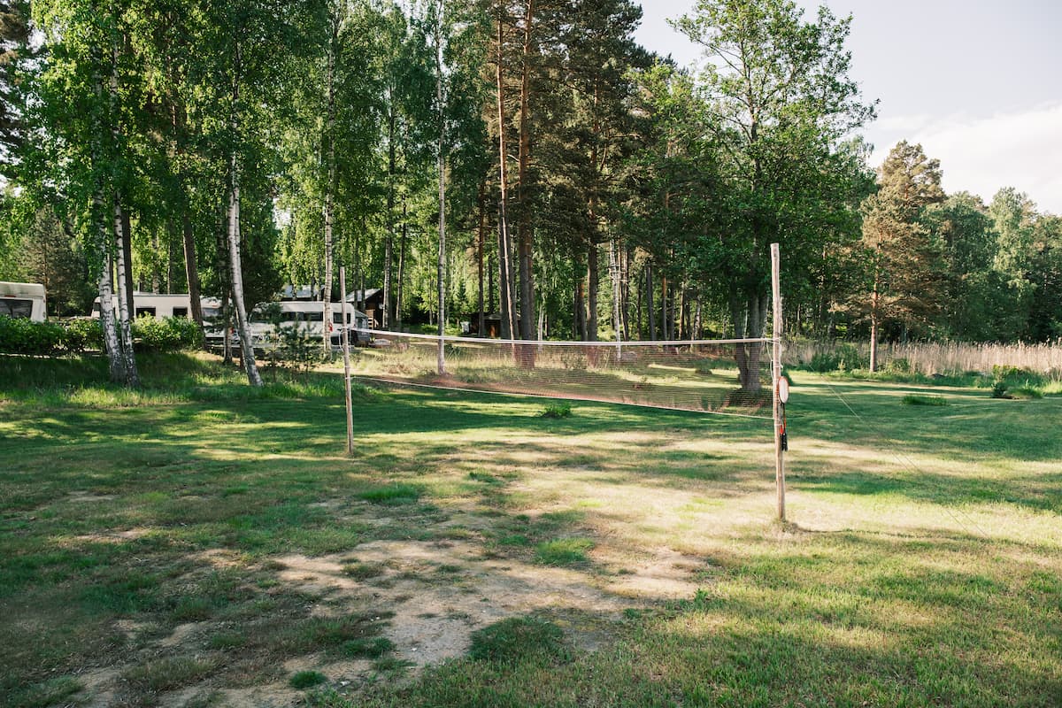 A grassy outdoor volleyball court with a net set up, surrounded by trees and parked camper vans in the background on a sunny day.