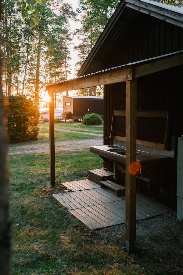 Wooden outdoor sink area attached to a dark cabin, with sunlight filtering through trees and another cabin in the background.