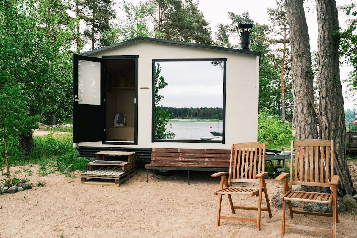 Small modern cabin with an open door, two wooden chairs on sandy ground, and a lake with trees in the background.