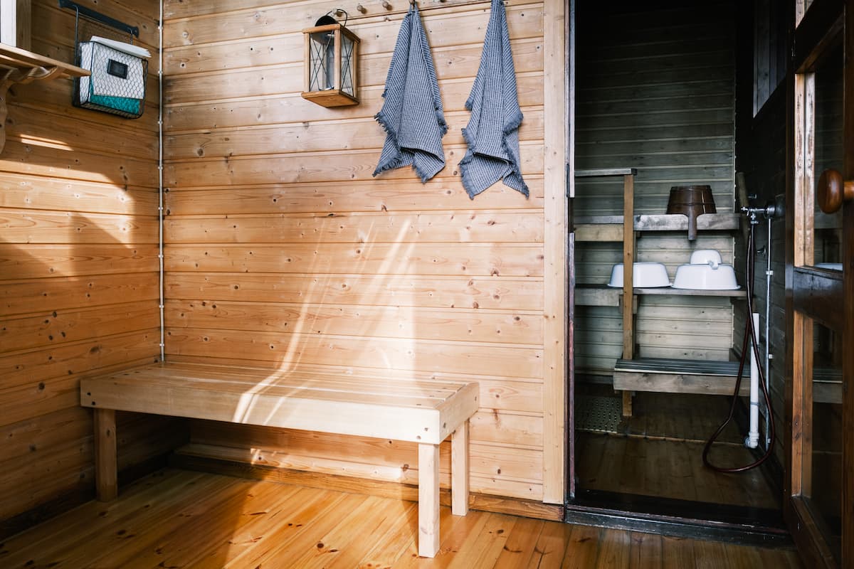 Wooden sauna interior with a bench, two grey towels hanging on hooks, a lantern on the wall, and a doorway leading to another sauna room with buckets and basins visible.