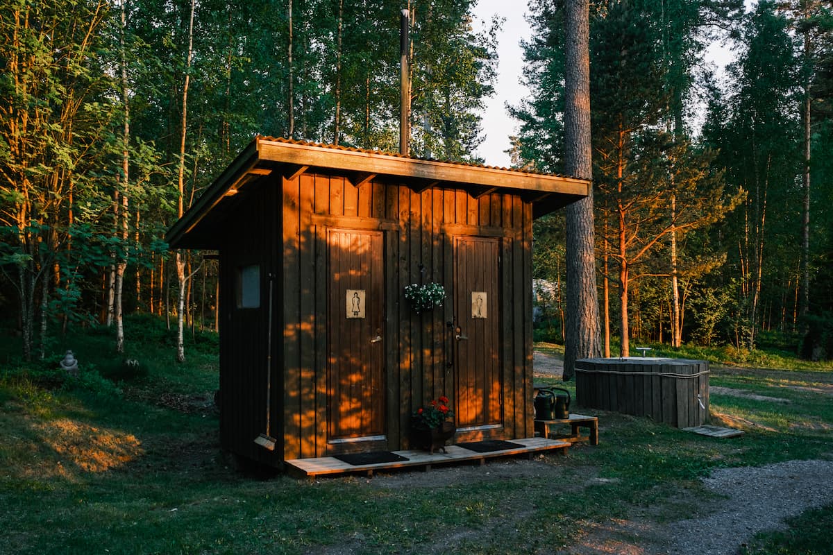 A small wooden outhouse with two doors stands among tall trees, lit by evening sunlight. A flower pot and plants are placed in front of the building.