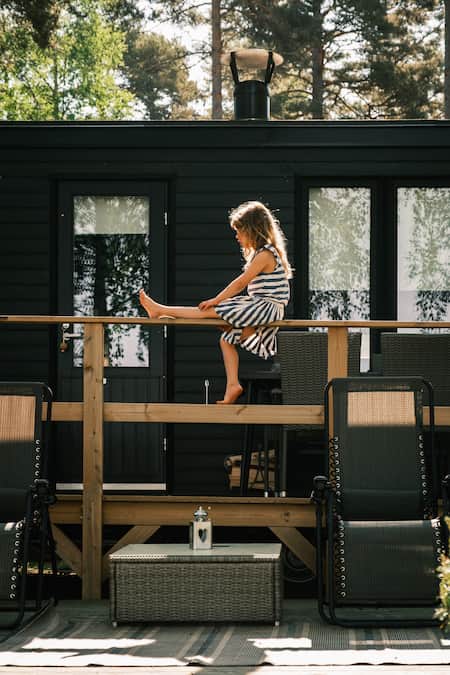 A young girl sits on a wooden railing in front of a dark house, stretching her legs whilst wearing a striped dress. Two sun loungers and a lantern are on the decking below.