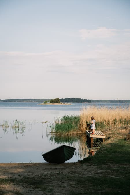 A child stands on a wooden jetty by a calm lake with reeds, an upturned boat onshore, and a small island visible in the distance under a clear sky.