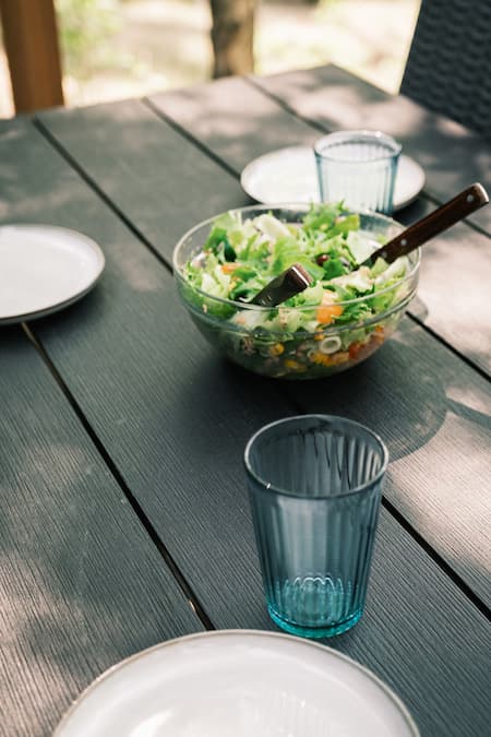 A glass bowl of salad with serving utensils on a dark wooden table, surrounded by empty plates and blue drinking glasses.