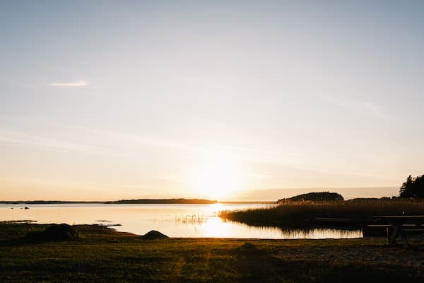 Sunset over a calm lake with grassy shore, a few reeds, and a wooden bench in the foreground.