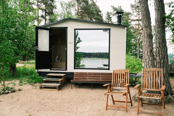 A small modern cabin with large window and open door sits on sandy ground near a lake, surrounded by trees, with two wooden chairs and a bench outside.