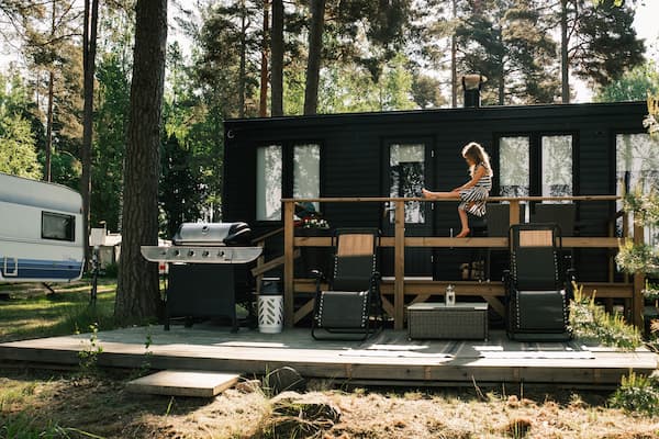 A child stands on the decking of a black mobile home in a wooded area, with outdoor chairs and a barbecue nearby. A white caravan is parked in the background.