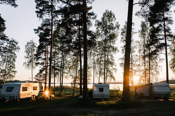 Three caravans are parked among tall trees at a lakeside campsite during sunset, with sunlight filtering through the branches.