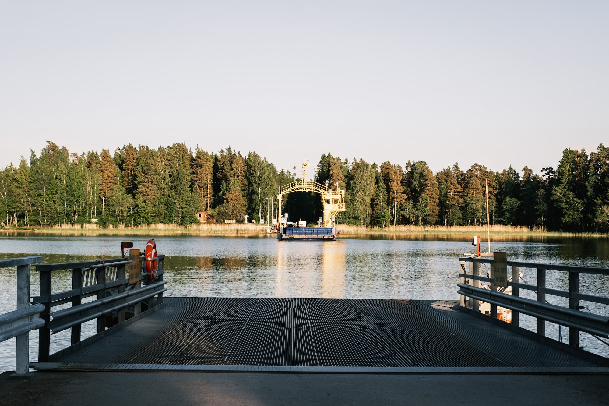 A ferry approaches a dock across calm water, surrounded by dense trees and a clear sky.