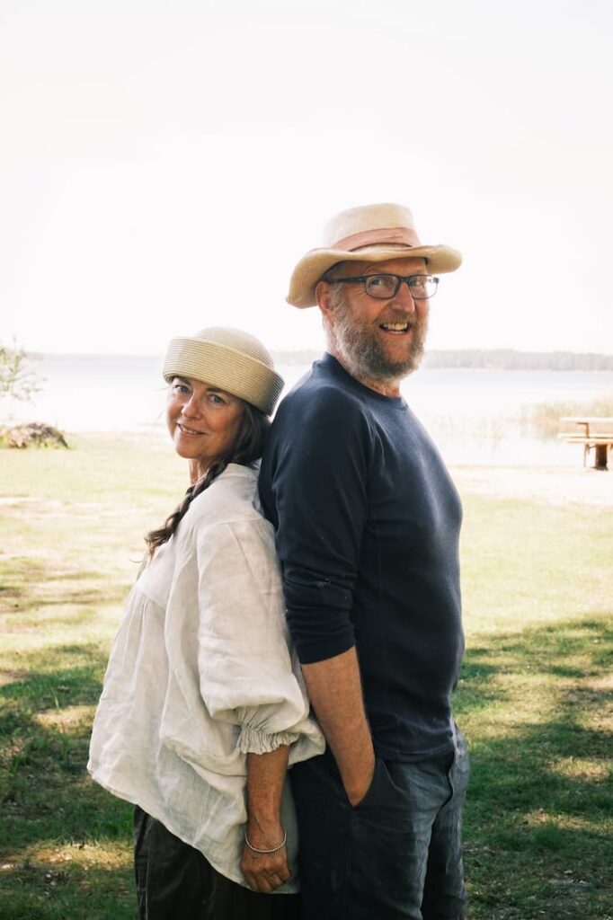 A woman and a man, both wearing hats, stand back to back outdoors on grass near water, smiling at the camera.