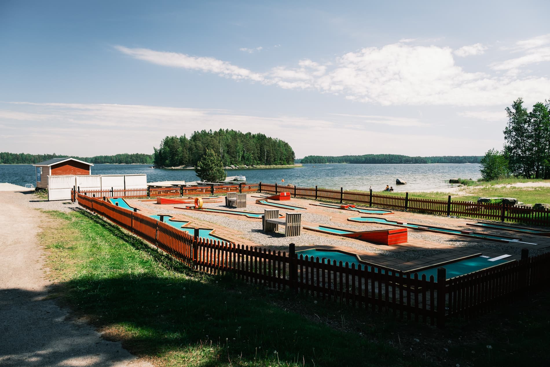 An outdoor mini-golf course with wooden fences is situated by a lake, surrounded by trees and a partly cloudy sky.