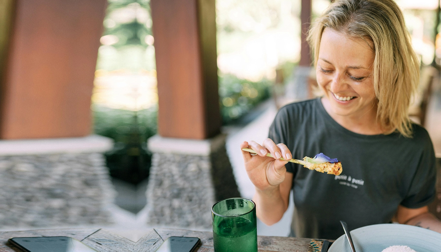 A woman sits at a table outdoors, smiling whilst holding food with chopsticks. A plate, a green glass, and a mobile phone are on the table.