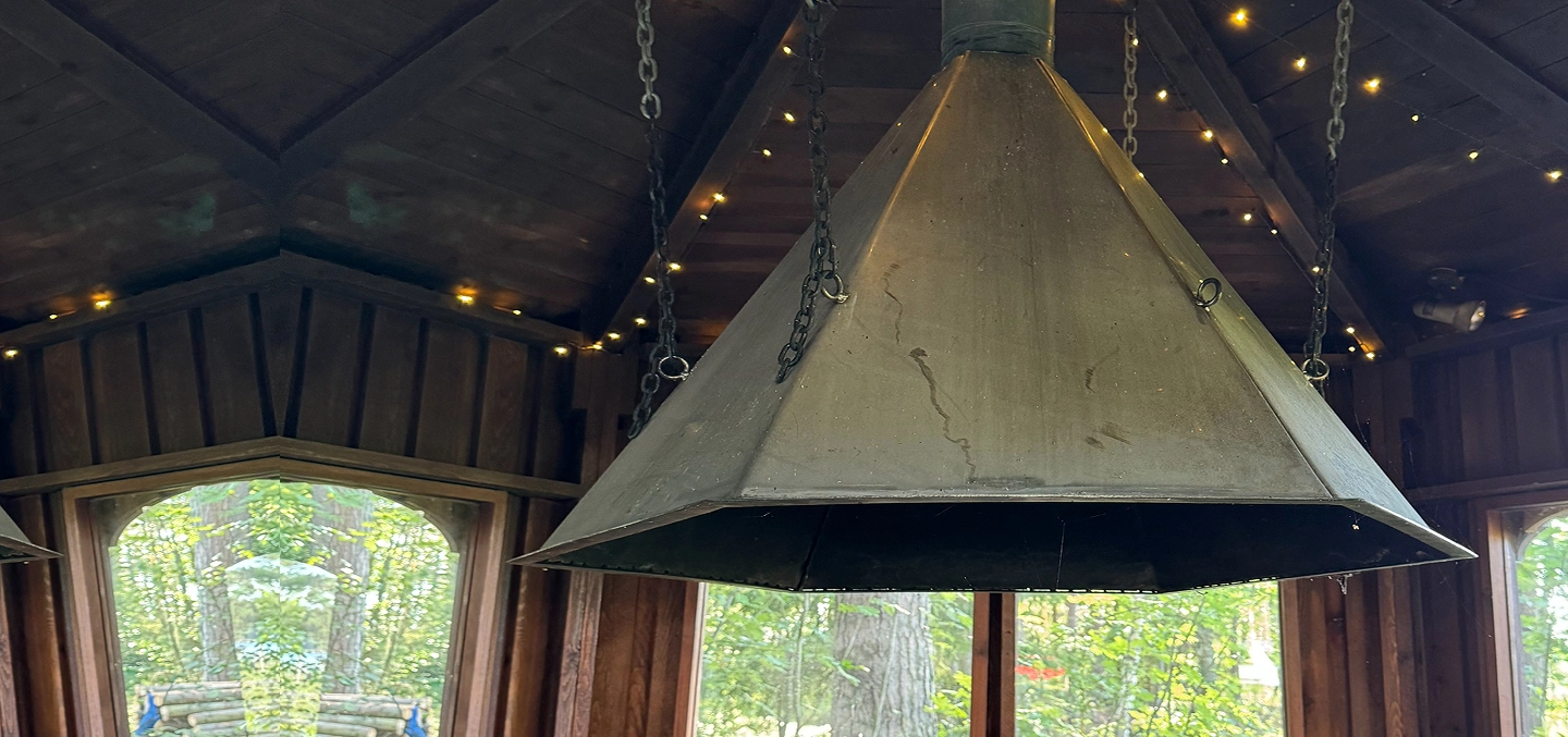 A large, metal, pyramid-shaped hood is suspended by chains from the ceiling in a wooden gazebo with windows overlooking trees. Small fairy lights are visible on the ceiling beams.